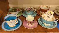 Photo showing six bone china teacups with matching saucers arranged on a shelf, showcasing different floral and decorative patterns and colors.