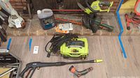 Two chainsaws (Ryobi cordless and Black & Decker corded) and Ryobi electric pressure washer with power cord and spray wand, laid out on floor.