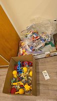 Overview of the lot showing multiple plastic bags with assorted Lego bricks and baseplates stacked near a cardboard box containing colorful plastic toy construction parts