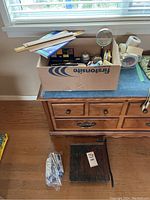 Box of assorted office supplies on a wooden dresser. Includes rulers, scissors, pens, tape, paper clips, and vintage paper cutter visible.