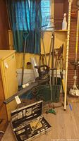 Gardening and outdoor tools including a shovel, rake, pole pruner, and gas leaf blower arranged near a window.