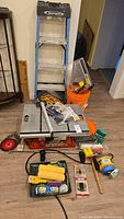 Full lot showing table saw base, ladder behind, orange bucket with assorted tools, paint rollers, paint tray and hand tools in front.
