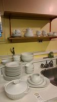 Overview of stacked plates, bowls, teapot, and other serving pieces displayed on countertop and shelf.