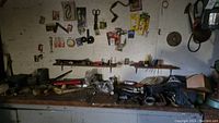 Wide view of a workbench surface with various scattered hand tools and hardware items including a vintage hand drill, clamps, pliers, and other miscellaneous tools.