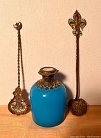 Two brass strainer spoons and turquoise glass bottle with brass stopper placed upright on a flat surface.