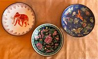 Three ceramic plates with brass frames laid out on a fabric surface, showing the elephant, floral, and butterfly designs.