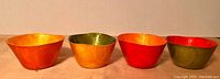 Four small dyed and lacquered Capiz shell bowls in varying colors (orange, green, red, olive green) arranged side by side on a surface.
