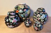 Front view of five vintage Chinese floral porcelain ginger jars, showcasing varying sizes and detailed hand-painted Millefleur Famille Noir style floral patterns on a black background.