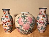 Photo showing all three Chinese porcelain vases together on a wooden surface against a beige background: two tall floral vases with butterflies flanking one round peach motif vase.