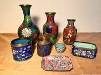 Photo showing seven cloisonné enamel pieces including three vases, three boxes/jars without lids, and one small cup, arranged on a wood surface against a plain background.