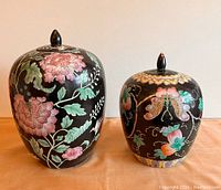 Two Chinese Famille Noir ginger jars with lids displayed side by side on a fabric surface, showing overall size and decorative details