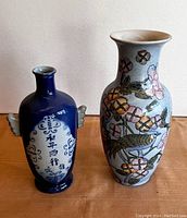 Two handpainted vases side by side on wooden surface. Left is a deep blue glazed ceramic bottle vase with white panel and Chinese script, small side handles, about 9 inches tall. Right is a taller handpainted porcelain vase with floral and bird design with pastel colors, about 10 inches tall.