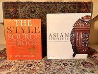 Two large format books placed on a patterned carpet. Left book is 'The Style Source Book' by Judith Miller with an orange cover, and right book is 'Asian Furniture' sourcebook edited by Peter Moss with a white cover and carved wooden furniture image.