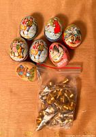 Overview photo of seven painted tin egg cases arranged on a table with a small plastic bag of brass goblets in front.