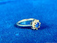 Side-angle view of the ring showing the blue spinel center and white topaz stones on a blue background.