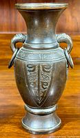 Front view of solid bronze urn vase showing dark patina, animal-head handles with rings, engraved pattern on body and neck.