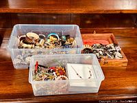 Two plastic bins and one wooden box on wooden surface containing assorted vintage and estate jewelry items such as rings, bangles, brooches, rhinestone pieces, silver-tone metal jewelry, and necklaces.