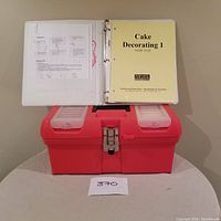 Photo of Cake Decorating 1 manual on top of a red plastic tool kit box with translucent trays inside the lid, placed on a round table.