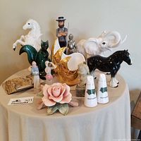 Photo showing large lot of figurines and decor items including ceramic horse, jade foo dog, golden vase, salt and pepper shakers, and marble ashtray on a round table.