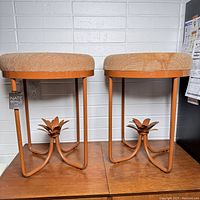 Full view of two orange metal framed stools with round beige cushioned seats and floral palm base detail, one still has original store tag visible