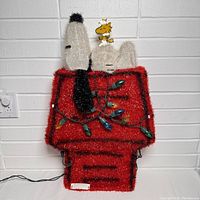 Photo showing Snoopy and Woodstock on a red doghouse with Christmas lights wrapped around, textured with tinsel material.