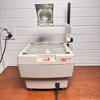 Front view of vintage 3M 1610 overhead projector showing glass projection surface, red slider control, and attached magnifying lens.