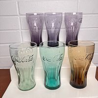 Front view of six colored vintage Coca Cola soda glasses showing three purple, one amber, one aqua blue, and one light green glass with embossed Coca Cola logo and contour shape.