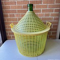 Front view of green glass demijohn bottle encased in yellow plastic carry basket with handles, showing bottle neck and lattice case.