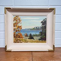 Front view of framed oil painting showing coastal landscape scene with autumn trees, water, cliffs, in off-white wooden frame with gold accents.