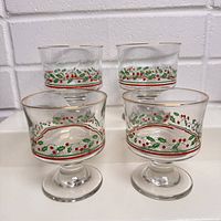 Four vintage juice glasses decorated with holly berry and green leaves, side view showing pattern and gold rim detail, set on a white surface with white brick background.