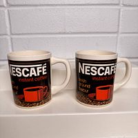 Two NESCAFÉ instant coffee mugs with black backgrounds, white NESCAFÉ text, red cup image, and coffee beans graphics.