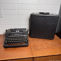 Smith Corona Sterling typewriter next to its black carrying case on wooden surface, showing front view and case closed.