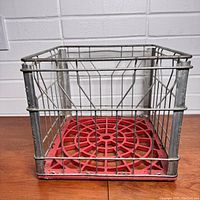 Square vintage wire milk crate with metal frame and red plastic base on wooden floor, front view