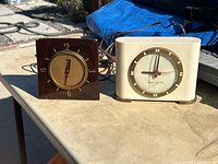 Two electric mantel clocks side by side on a table outdoors, showing one dark brown square clock and one cream-colored rectangular clock.
