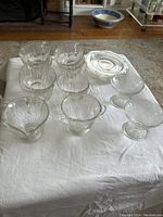 Seven clear patterned coffee cups, five matching saucers, one creamer, one sugar bowl, and two footed sherbet glasses arranged on a white cloth.