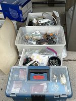 Plastic bins filled with assorted hardware items including nuts, bolts, and miscellaneous fittings placed on pavement.