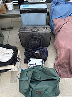 Photo showing assortment of luggage and backpacks on a concrete floor, including a large blue suitcase, grey fabric suitcase with locks, black and navy backpacks, blue and white soft bag, and green duffel bag.