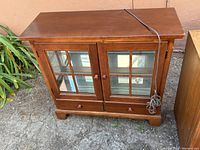 Front view of cherry wood display cabinet with closed glass doors, two drawers at the base, and electrical cord for internal lighting.