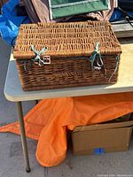 Closed wicker picnic basket on a table with leather straps and buckles, green leather fastening ribbons.