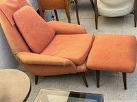 Three-quarter side view of orange lounge chair with ottoman showing leg finish and upholstery wear.