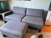 Gray modern sofa with two seat cushions, two back cushions, and wooden legs visible. Includes matching ottoman in front.