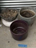 Top view of round ceramic pot with metal scrollwork rim and white base, filled with dry leaves and twigs, next to large grey cylindrical pot with soil inside and smaller purple pot partly visible