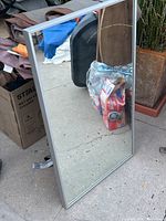 Full view of a new rectangular mirror with a silver frame standing upright outdoors, showing the mirror surface and surrounding area.