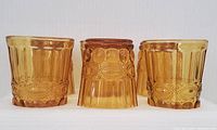 Front view shows six amber glass tumblers standing upright, highlighting the textured vertical facets and embossed decorative band.