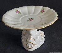 Top and side view of a white porcelain scalloped dish with pink floral motifs and gold edges on dolphin head pedestal base.