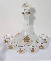 Full set view showing one decanter with stopper and six matching shot glasses arranged around it, all decorated with gilded ship and gold accents