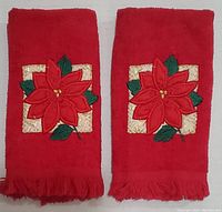 Two red velvety hand towels side by side, each with a poinsettia applique centered on a gold square and fringe on one edge