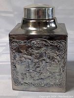 Front view of square silver plated vessel with embossed pastoral scene featuring gazebo and figures in garden.