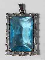Front view of rectangular blue glass pendant surrounded by clear baguette rhinestones in silver-tone setting.