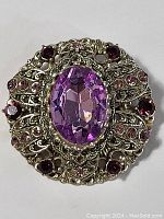 Front-facing view of vintage brooch showing large oval central purple gemstone with surrounding smaller rhinestones and ornate silver-tone metal filigree.